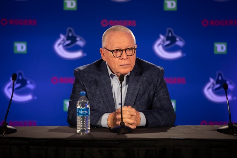 A man in a dark suit and white open-collared shirt sits at a desk in front of a microphone with a plastic water bottle beside him, waiting for questions. Behind him is the Vancouver Canucks large-C-and-orca logo on a blue background.