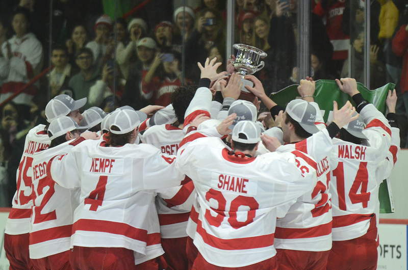 Hockey players hoist a trophy at the side of a hockey rink with fans watching and cheering through the glass