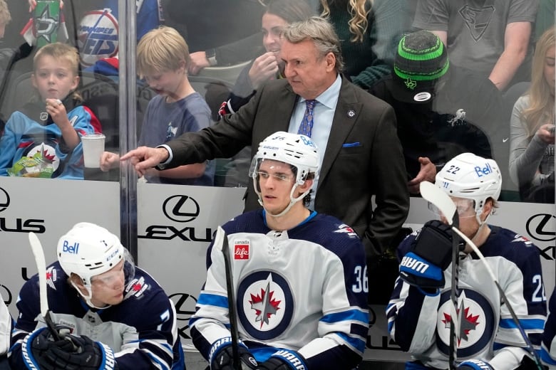 A man in a suit points toward the ice while standing behind the Winnipeg Jets bench, where three players are seated in front of him.