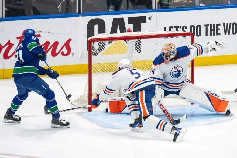 A hockey player scores past an outstretched goalie, as a defender tries to block his shot.